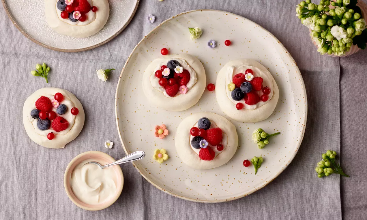 Na svijetlom tanjuru Mini-Pavlova bijele boje s hrskavim rubovima, na sredini svježe crvene maline i ribiz, plave borovnice