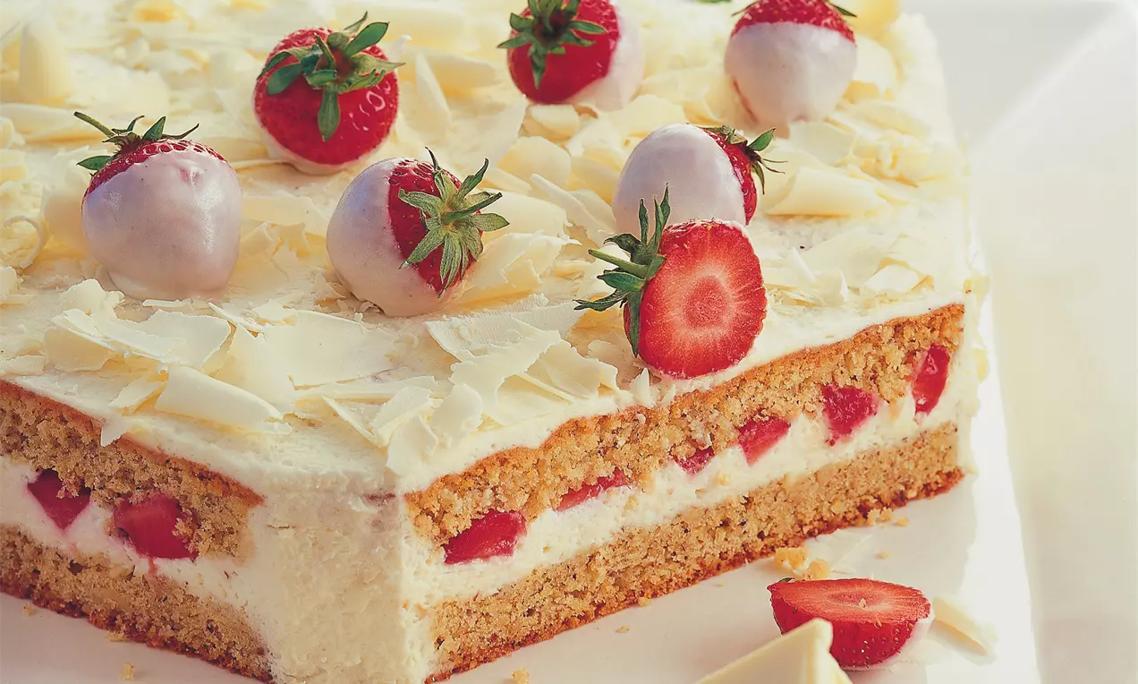 Torta pravokutnog oblika s bijelim slojem šlaga, ukrašena jagodama i bijelom čokoladom na svijetlom tanjuru