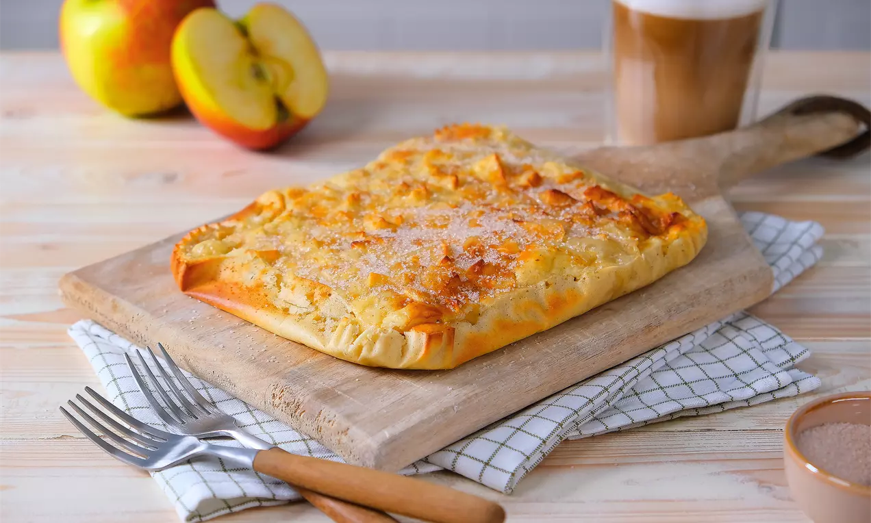 Zlatnožuti pravokutni kolač s hrskavom korom posut kristal šećerom na drvenoj dasci, uz jabuke, latte i pribor za jelo.