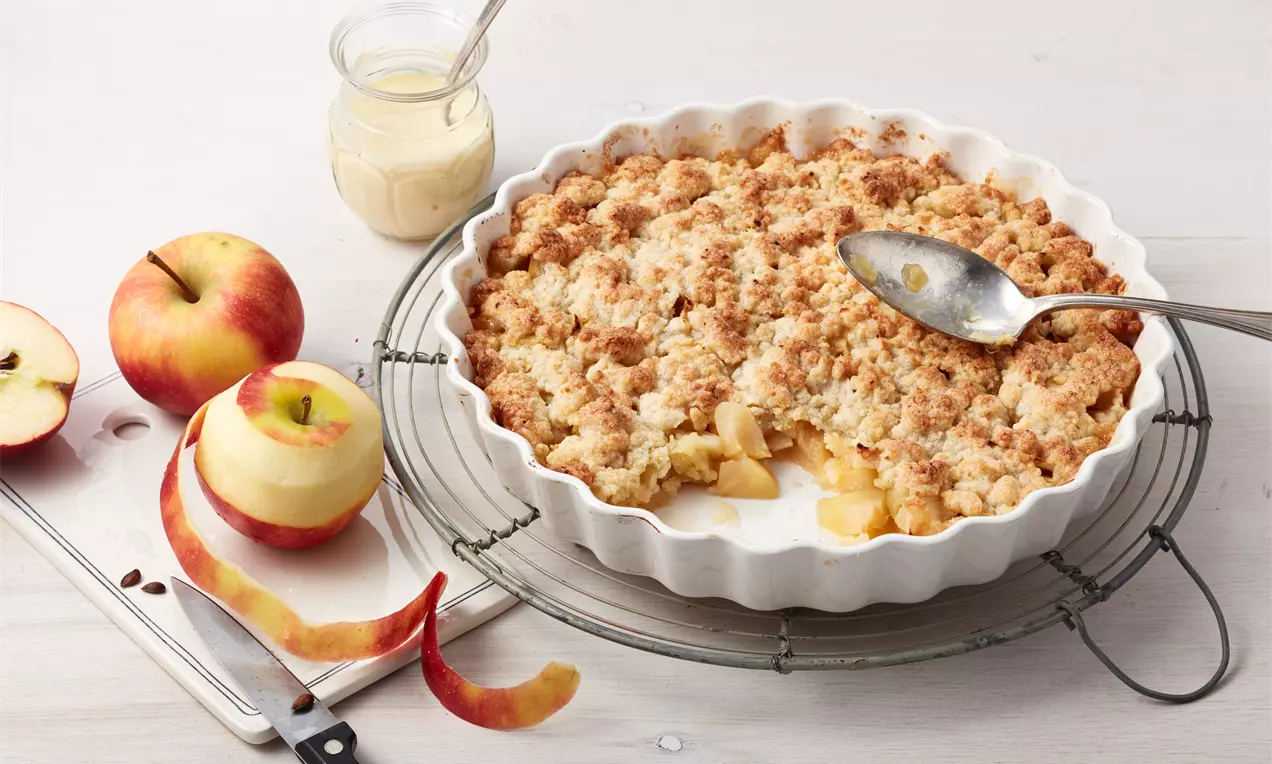 Zlatnosmeđi crumble s neravnom površinom na bijelom rebrastom kalupu, uz jabuke, koru i kremu u staklenci na drvenoj podlozi