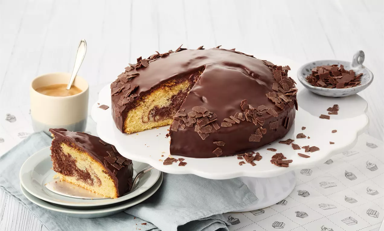 Torta s glatkom tamnom čokoladnom glazurom, posuta čokoladnim strugotinama, posluženom na bijelom stalku uz šalicu kave i tanjurić