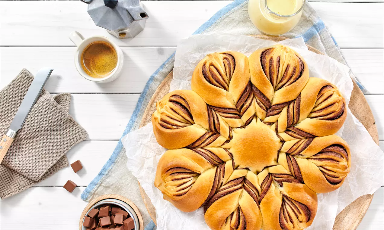 Zvjezdasti oblik zlatnosmeđih buchtli s tamnim spiralnim uzorkom od čokolade na papiru za pečenje, okružen šalicom kave i komadićima čokolade