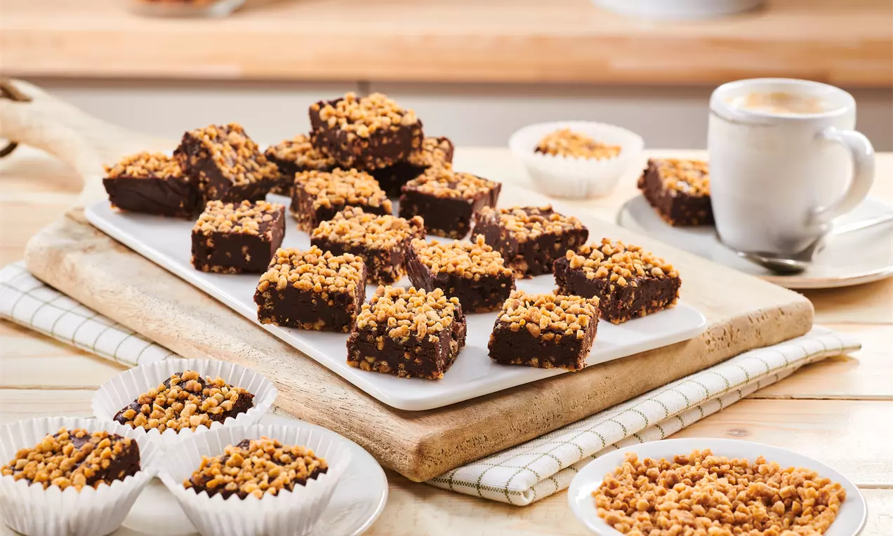 Čokoladne kocke tamnosmeđe boje s hrskavim zlatnim posipom na vrhu, posložene na bijelom pladnju uz šalice kave i zdjelice s posipom