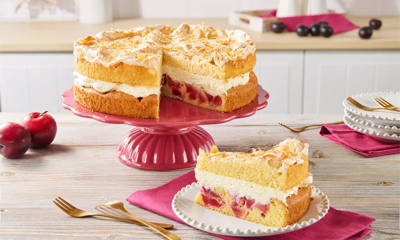 Slojevita torta na crvenom stalku s biskvitom, kremom, komadićima šljiva i zlatnosmeđim beze slojem, uz tanjur sa šnitom na stolu.