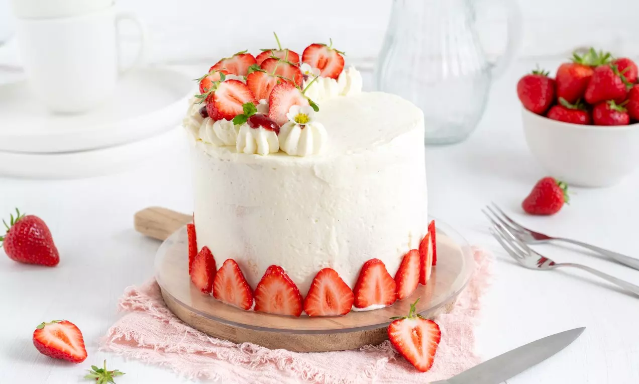 Okrugla torta svjetlo bijele glazure ukrašena šlagom i svježim jagodama na vrhu, s narezanim jagodama uz rub, na drvenom podlošku