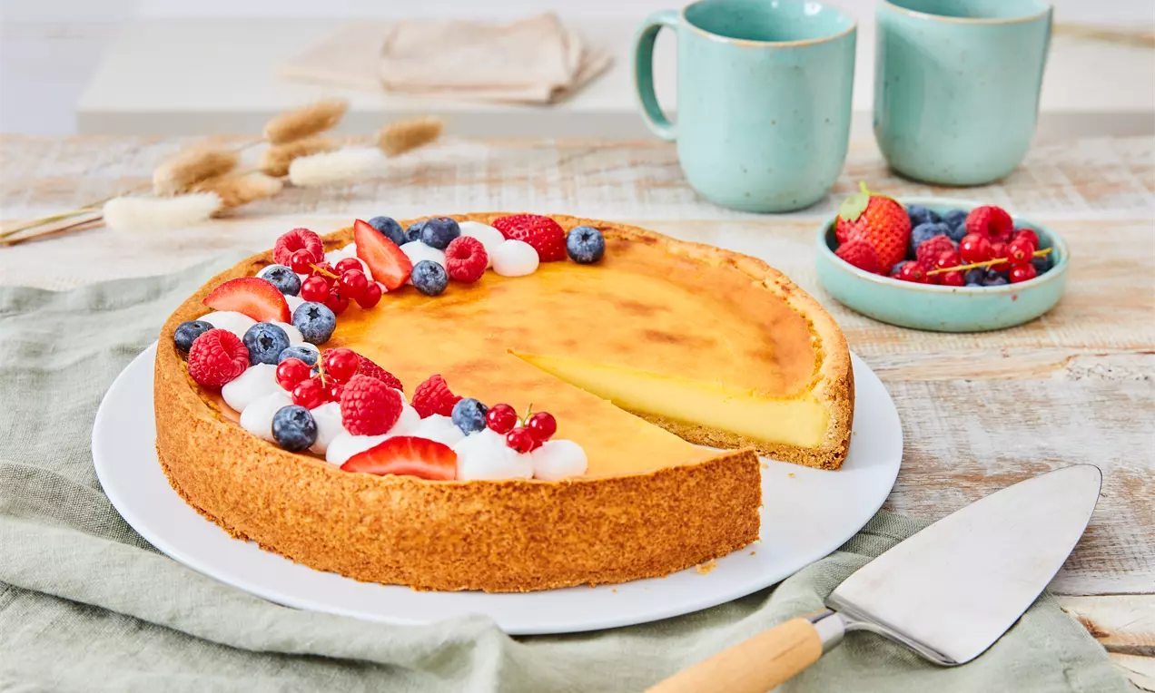 Vegan cheesecake sa zlatno-smeđim punjenjem, okružen dekorativnim vrpcama šlaga, svježim bobičastim voćem i hrskavom podlogom na bijelom tanjuru