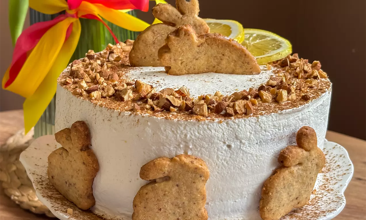 Torta bijele površine ukrašena zlatnosmeđim zec kolačićima, mrvicama oraha i kriškama limuna na bijelom stalku