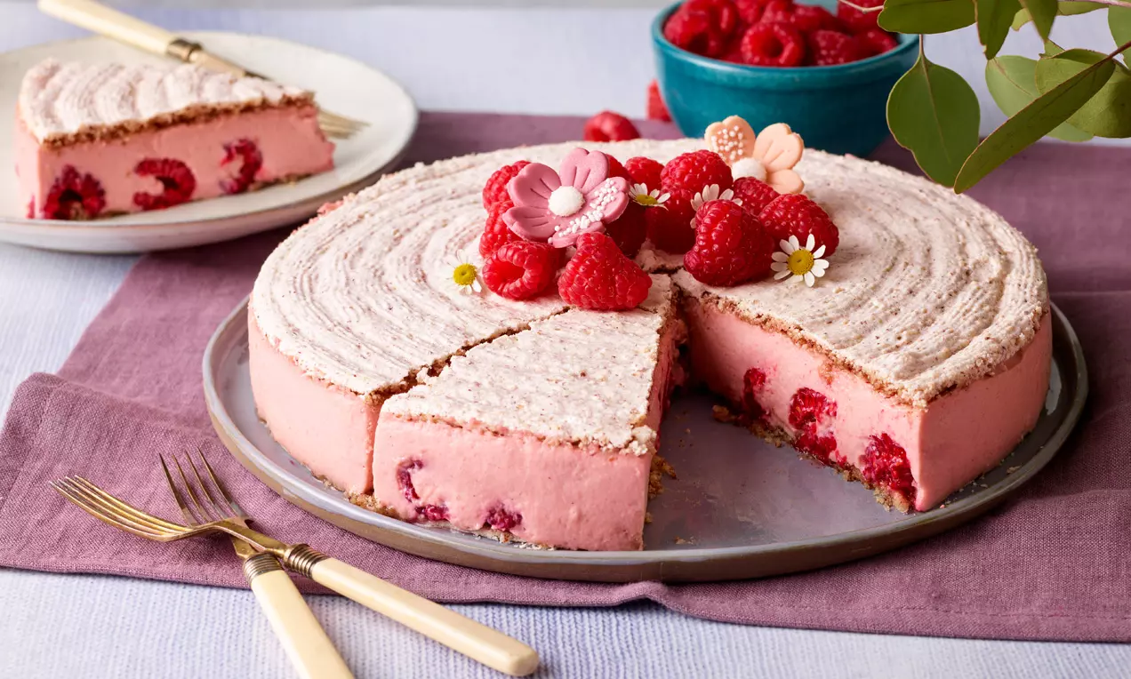 Okrugla torta s kremom svijetloružičaste boje, ukrašena malinama, jestivim cvjetićima i poslužena na plosnatom tanjuru