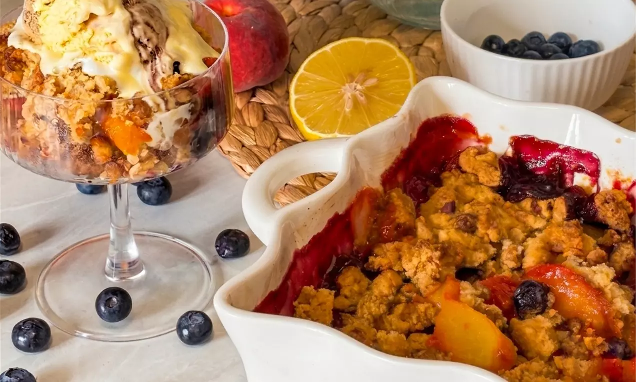 Voćni crumble s tamnoljubičastim bobicama i zlatnim breskvama u keramičkoj posudi, uz čašu s crumbleom, sladoledom i voćem