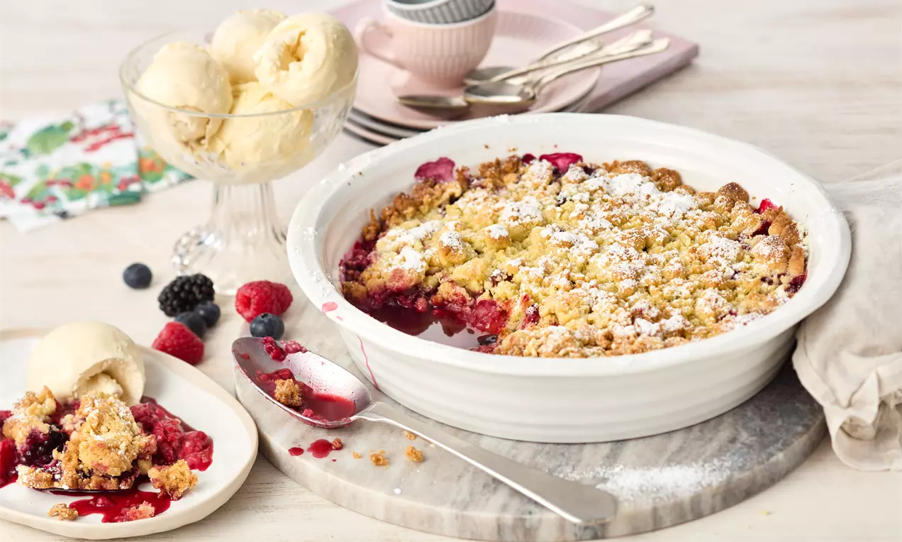 Pečeni voćni crumble u bijeloj keramičkoj posudi, s mješavinom crvenih bobica, zlatnosmeđim posipom i tragovima šećera u prahu.