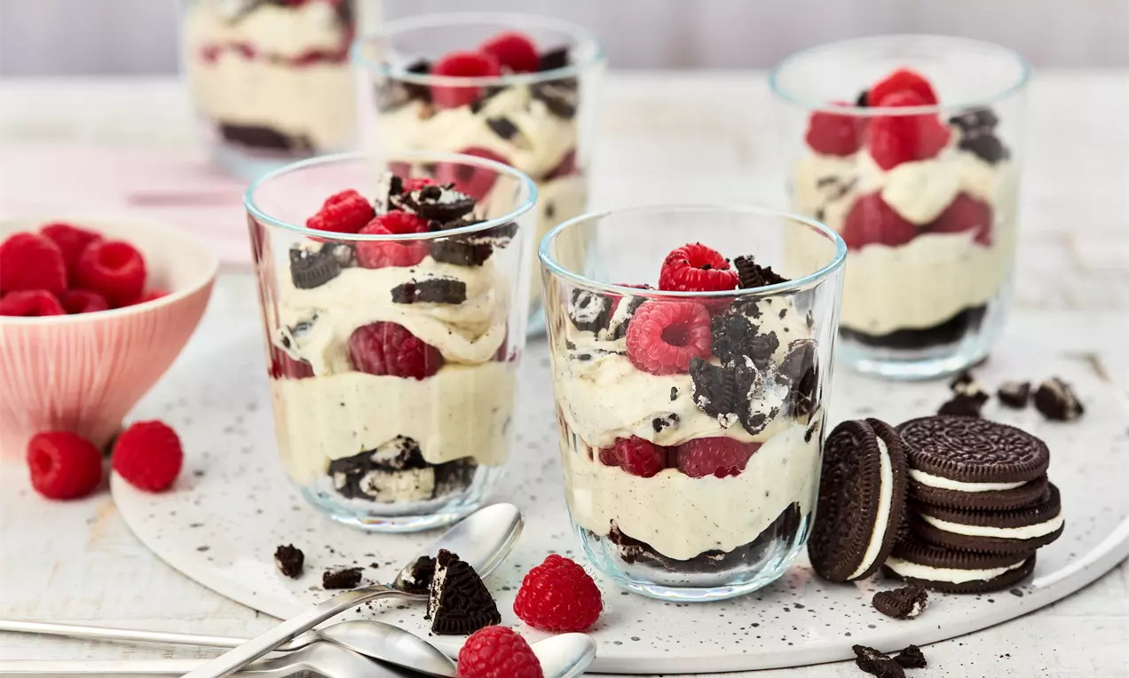 U prozirnim čašama slojevi svijetle kreme, svježih crvenih malina i tamnih komadića Oreo keksa na bijelom podlošku sa žlicama