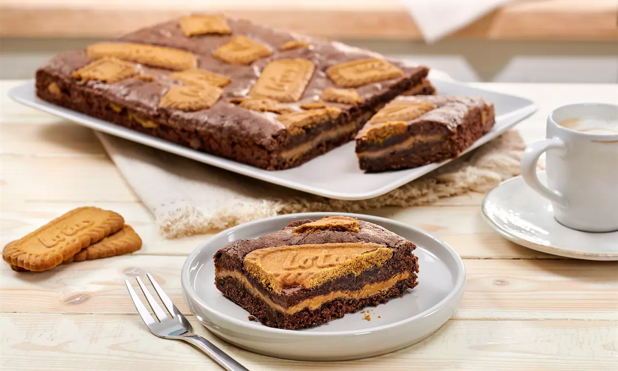 Čokoladni brownie s mekim slojem, ukrašen hrskavim Lotus keksima na vrhu, poslužen na bijelom tanjuru uz vilicu na drvenoj podlozi