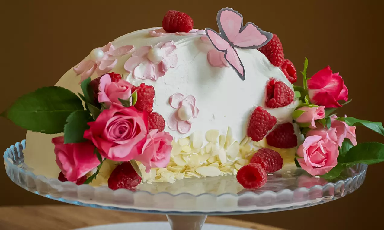 Bijela kupolasta torta ukrašena svježim malinama, ružičastim ružama, papirnatom leptir dekoracijom i listićima badema na staklenom postolju