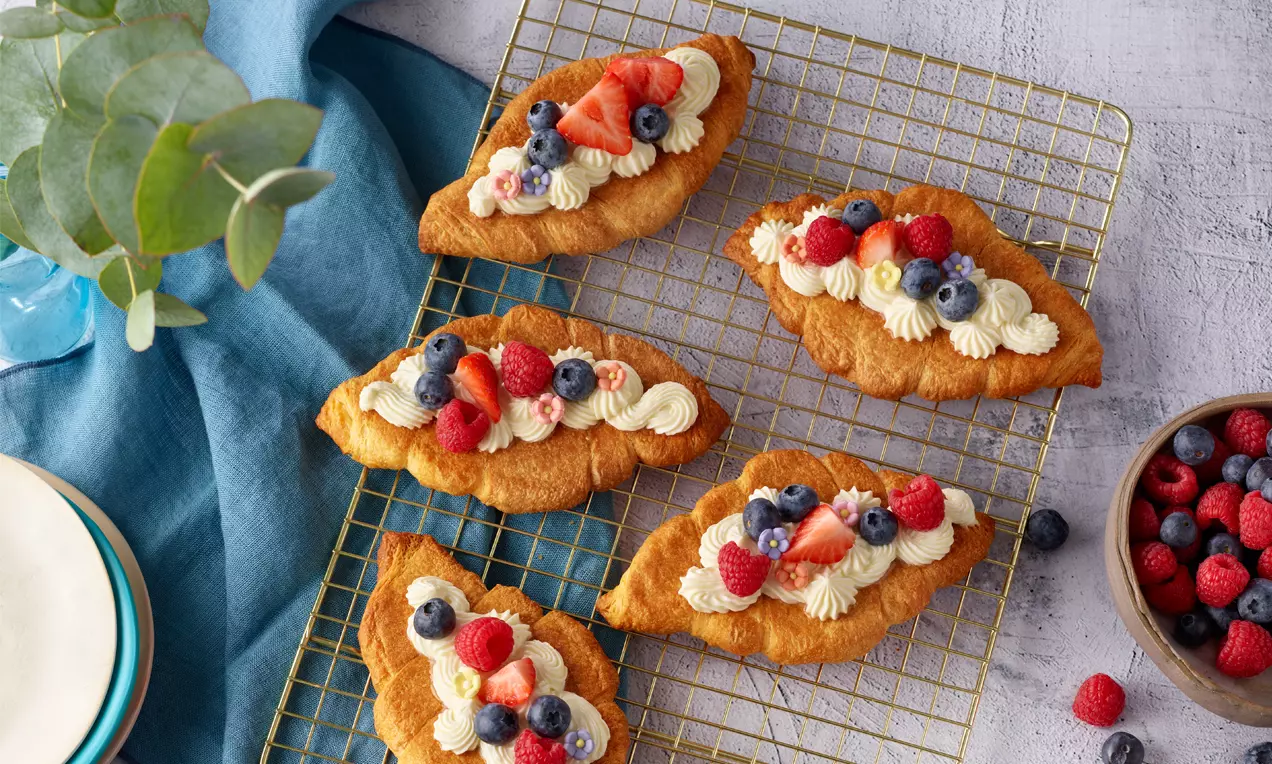 Zlatnosmeđi kroasani tankog lista, ovalnog oblika, na rešetki, prekriveni bijelom kremom i ukrašeni svježim borovnicama, malinama i jagodama