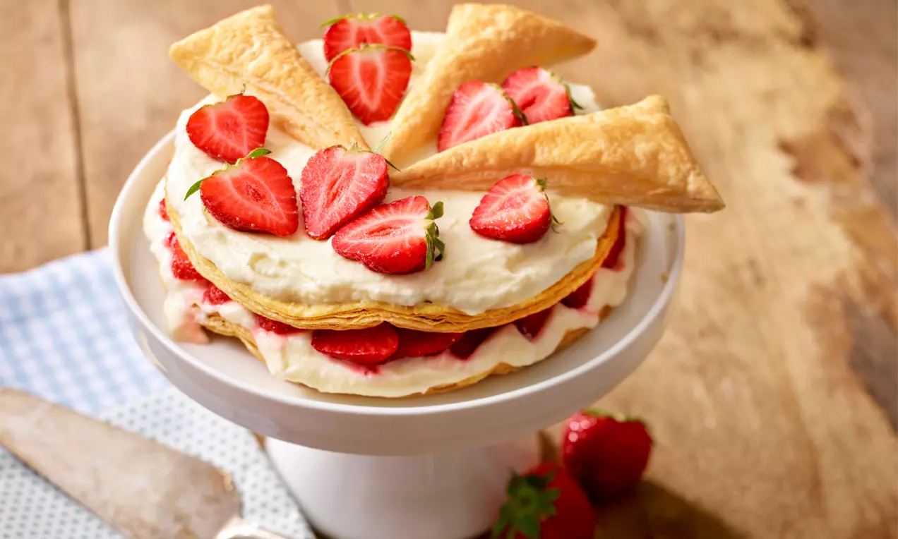 Torta od lisnatog tijesta svijetlozlatne boje, slojevita s kremom i svježim jagodama, ukrašena trokutima tijesta na bijelom stalku