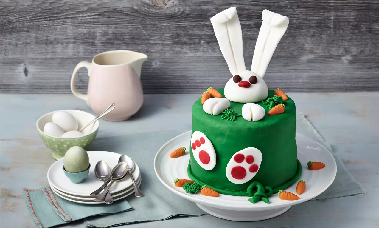 Torta s glatkom zelenom glazurom, ukrašena bijelim ušima, njuškom i šapicama zečića, uz male narančaste mrkve na bijelom pladnju