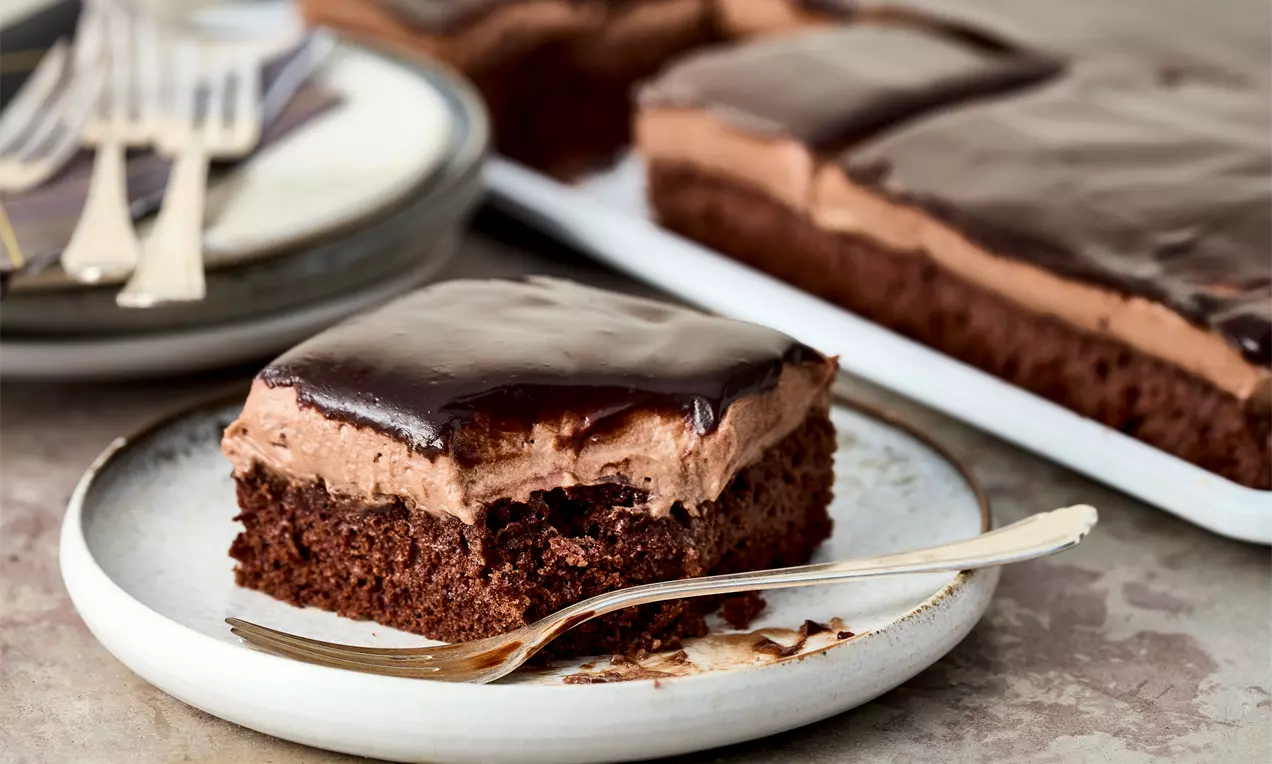 Čokoladni kolač s tamnim sjajnim ganache preljevom, slojem pjenaste čokoladne kreme i mekanom biskvitnom podlogom na keramičkom tanjuru.