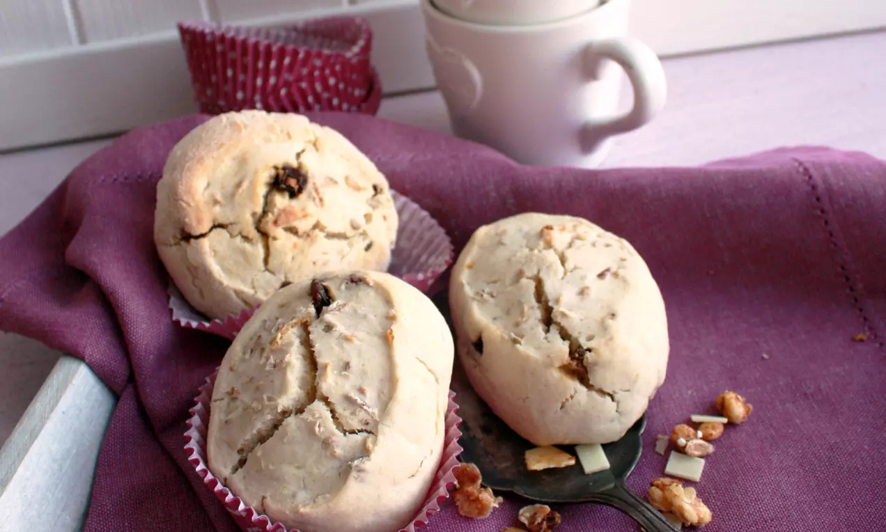 Svijetlosmeđi okrugli scones s pukotinama na površini, posluženi na ljubičastom ubrusu u papirnatim košaricama uz bijelu šalicu