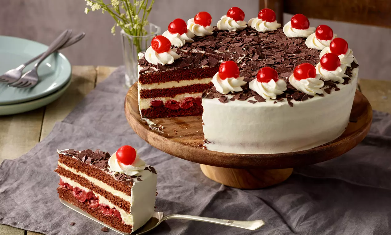 Torta prekrivena bijelom kremom, čokoladnim strugotinama i šlagom, ukrašena crvenim višnjama, s tanjurom i izrezanim komadom na platnenom stolnjaku