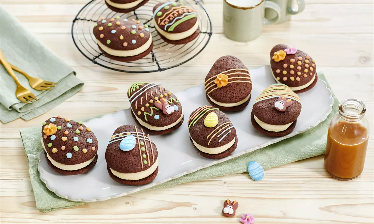 Tamnosmeđi kolači u obliku jaja s bijelim kremastim punjenjem, ukrašeni šarenim uzorcima i motivima na bijelom pladnju na drvenom stolu