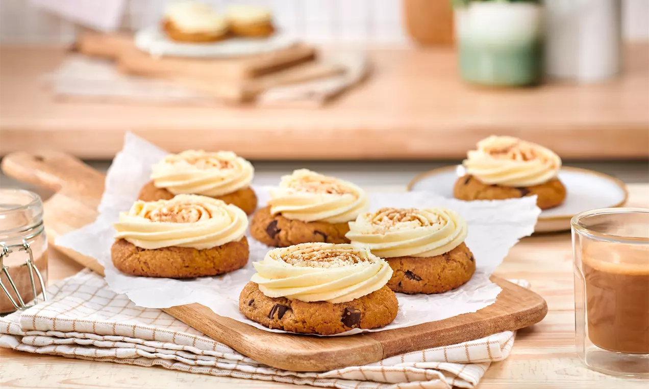 Na drvenoj plati, zlaćano smeđi keksi s hrskavim rubovima, ukrašeni svijetlim kremastim zavojima i mrvicama, okruženi toplim detaljima kuhinje