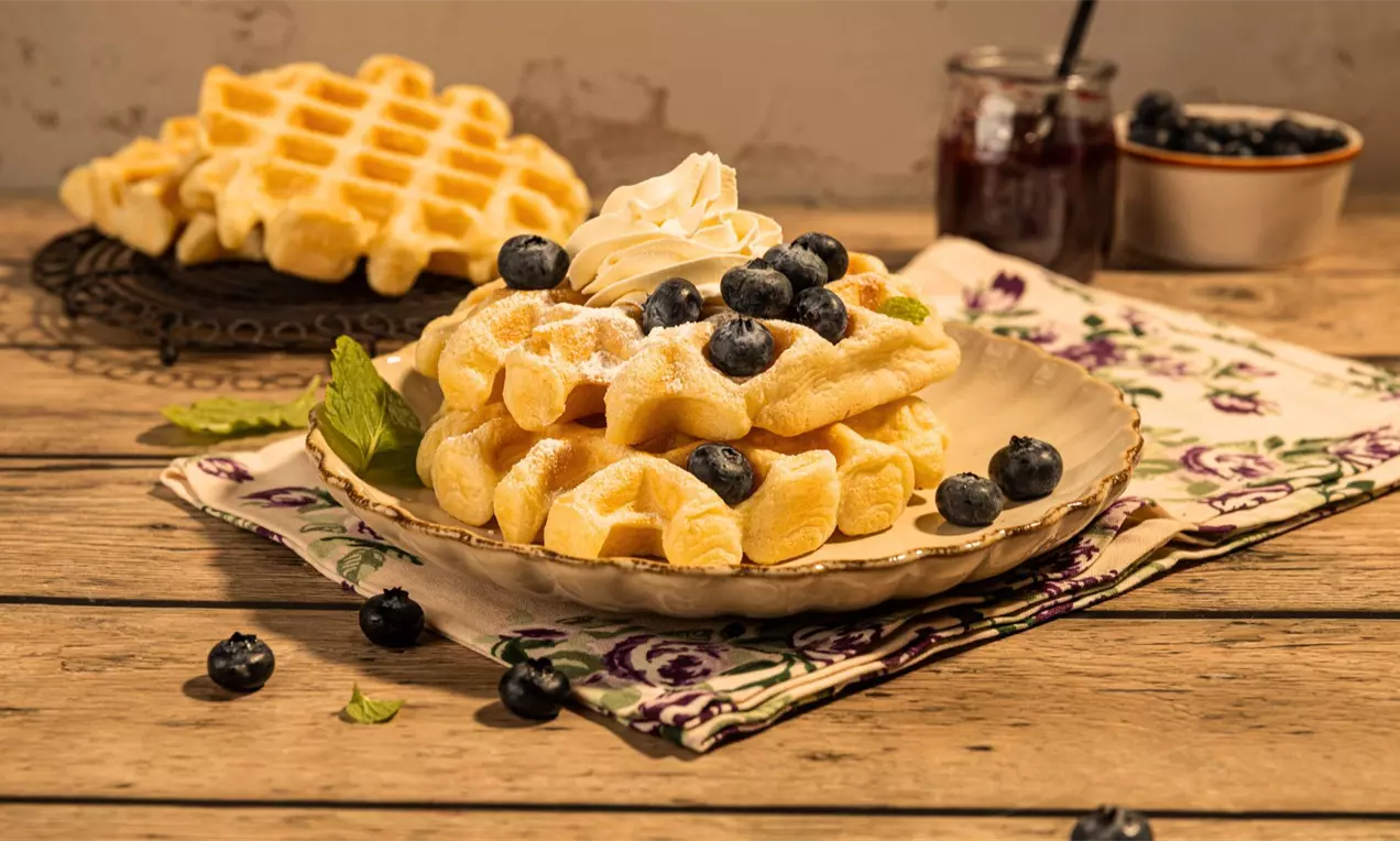 Zlatnosmeđi vafli poslagani na plavom tanjuru s bijelim uzorkom, ukrašeni tučenim vrhnjem, svježim borovnicama i šećerom u prahu