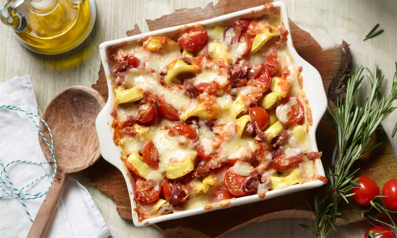 Pečeni tortelloni u bijeloj posudi, prekriveni rastopljenim sirom, kriškama cherry rajčica, par pršuta i komadićima tikvica