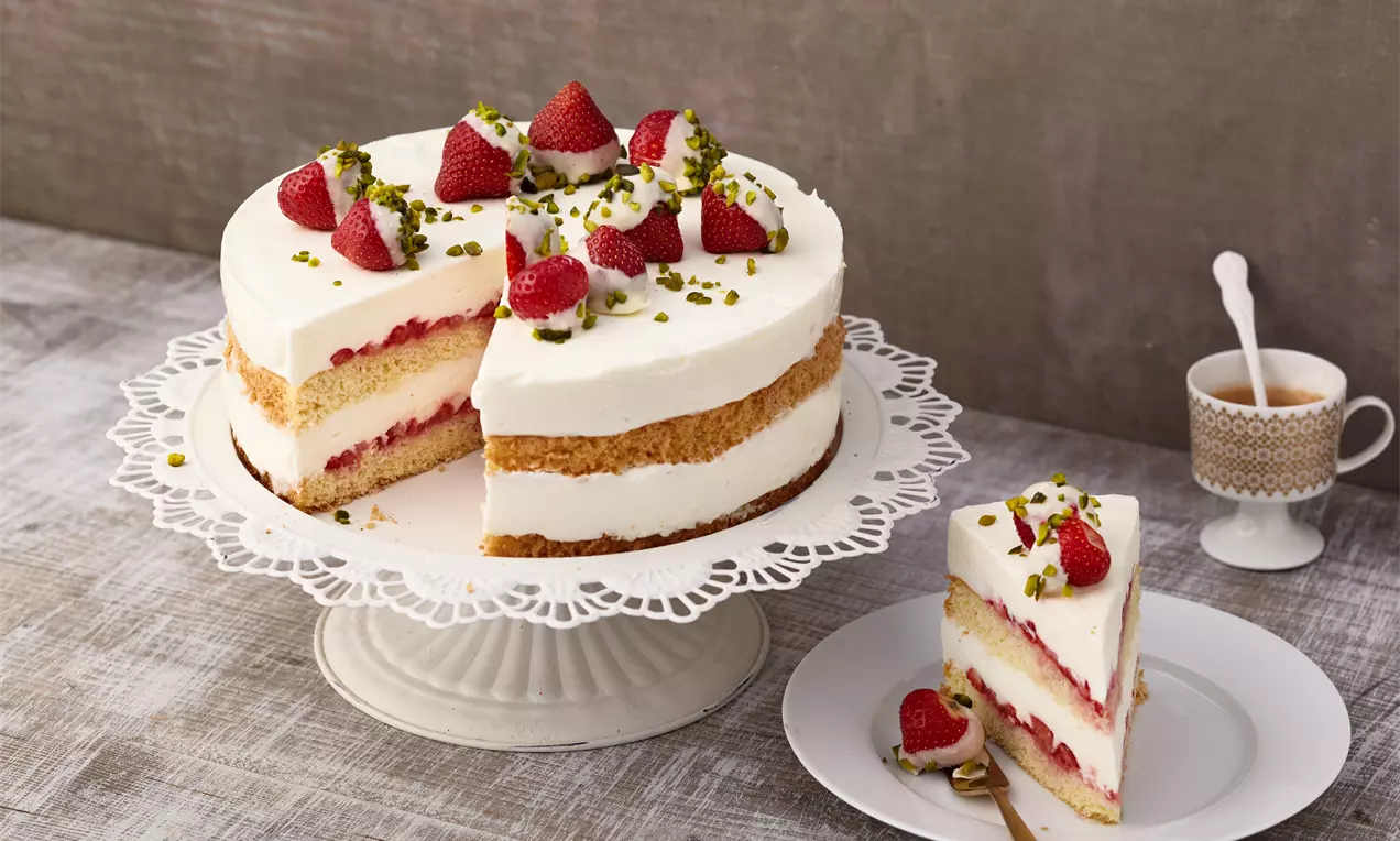 Torta s bijelom kremom, slojevima biskvita i jagodama na čipkastom postolju, ukrašena pistacijama i jagodama s kriškom na tanjuru