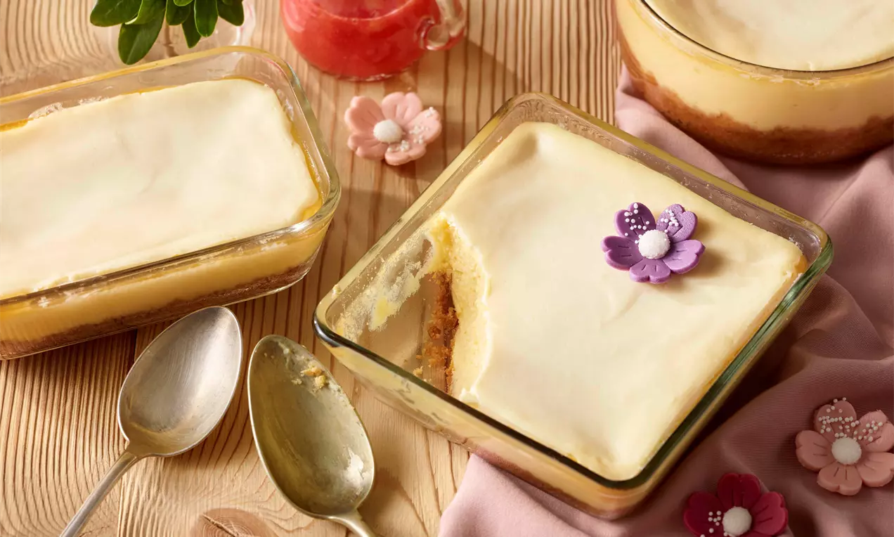 Kremasti New York cheesecake blijedožute boje, u staklenoj posudi s cvjetnim ukrasima, djelomično zagrizan, na drvenom stolu.