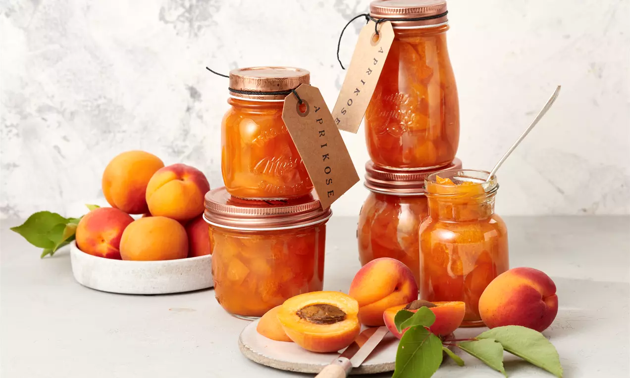 Staklenke narančaste marmelade od marelica različitog oblika, s čepovima i tagovima, uz svježe marelice na svijetlom podlošku