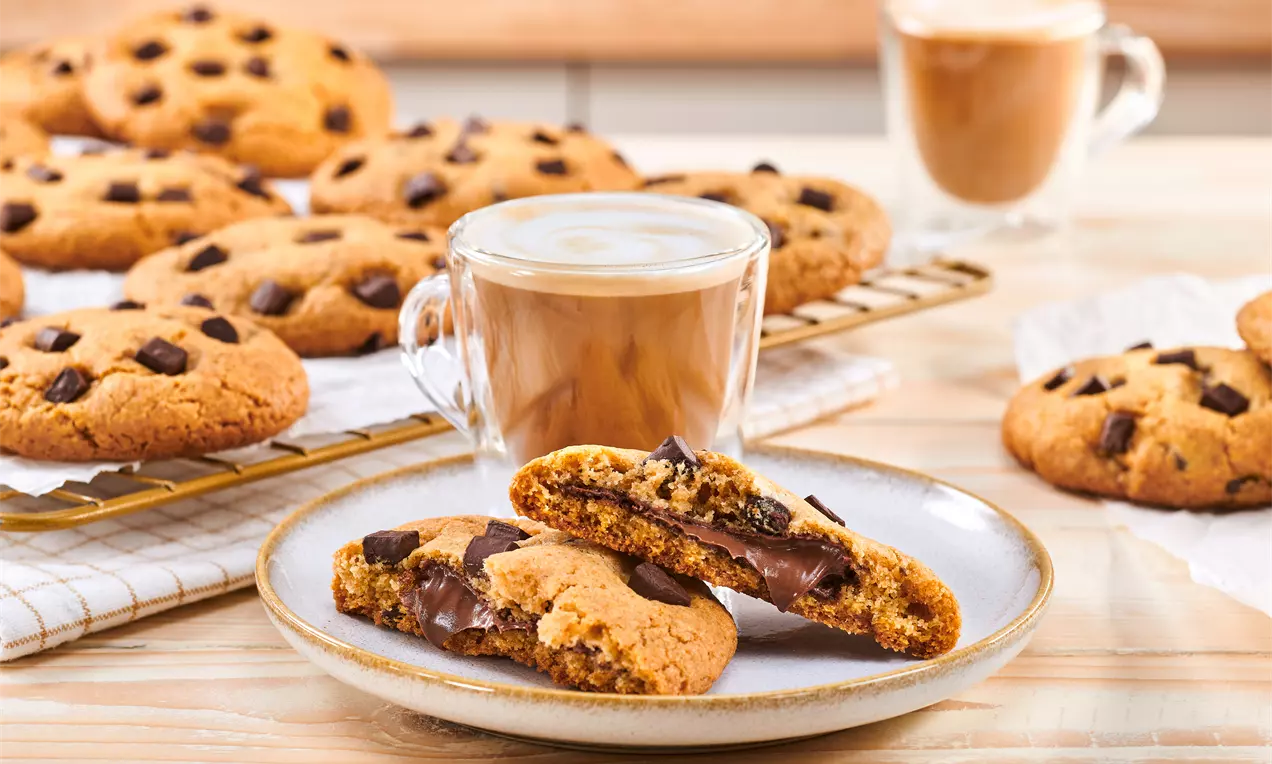 Zlatnosmeđi kolačići s komadićima čokolade, jedan prepolovljen otkrivajući Nutella punjenje, na tanjuru uz šalicu kave i rešetku s kolačićima