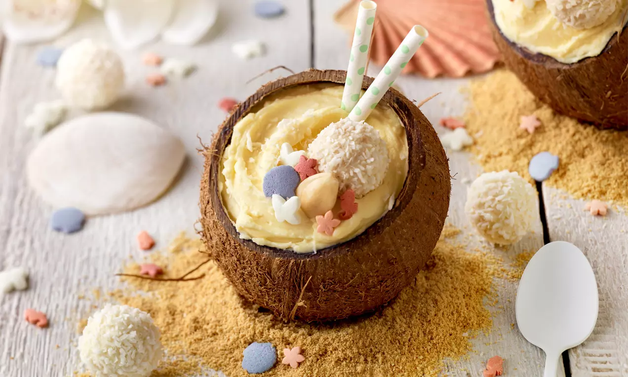 Svijetložuta kremasta mousse poslužena u polovici kokosova oraha, ukrašena bijelim Raffaello kuglicama, pastelnim dekoracijama i slamkama