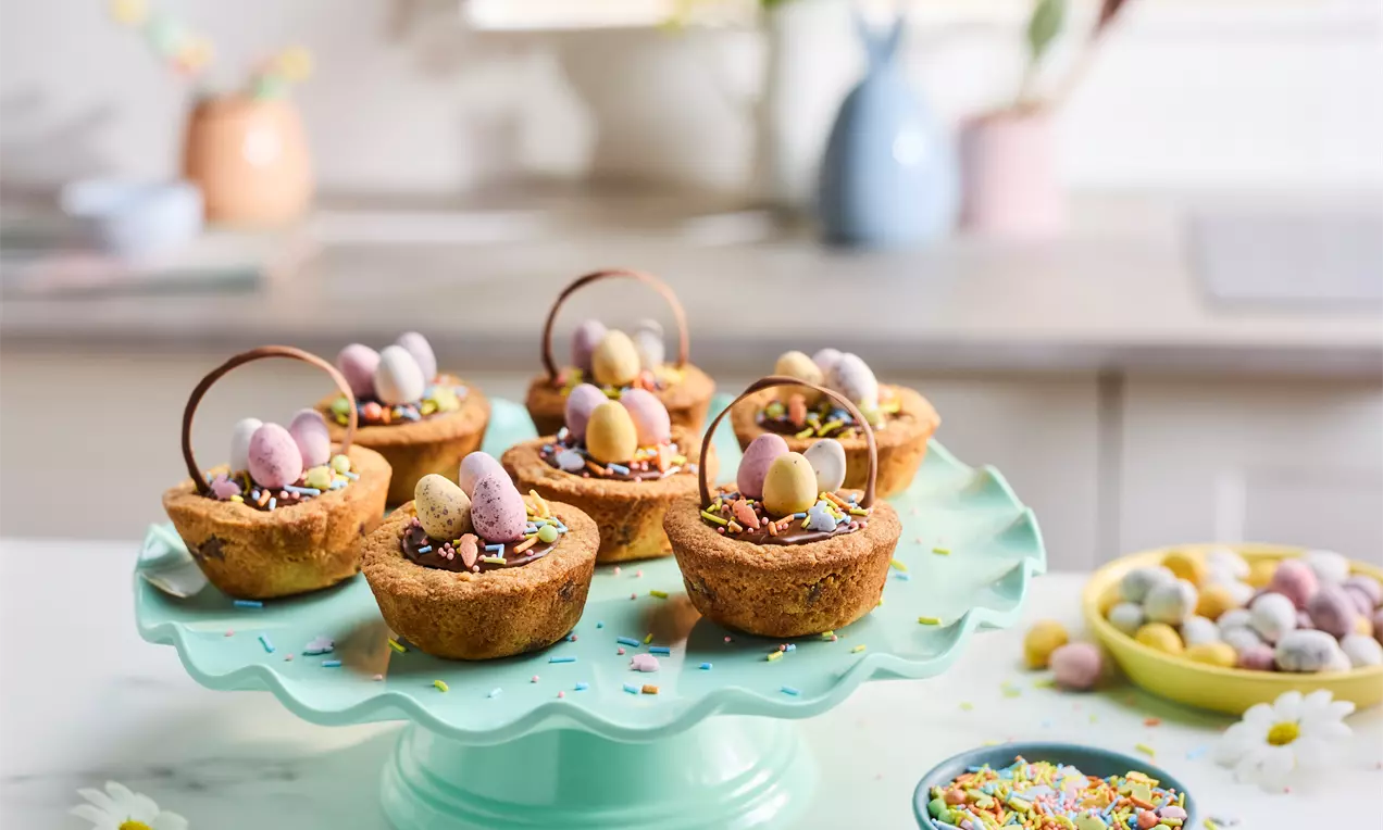 Mini košarice od zlatnosmeđeg tijesta na zelenom stalku, ispunjene šarenim jajima, čokoladom i šarenim mrvicama, ukrašene čokoladnim drškama