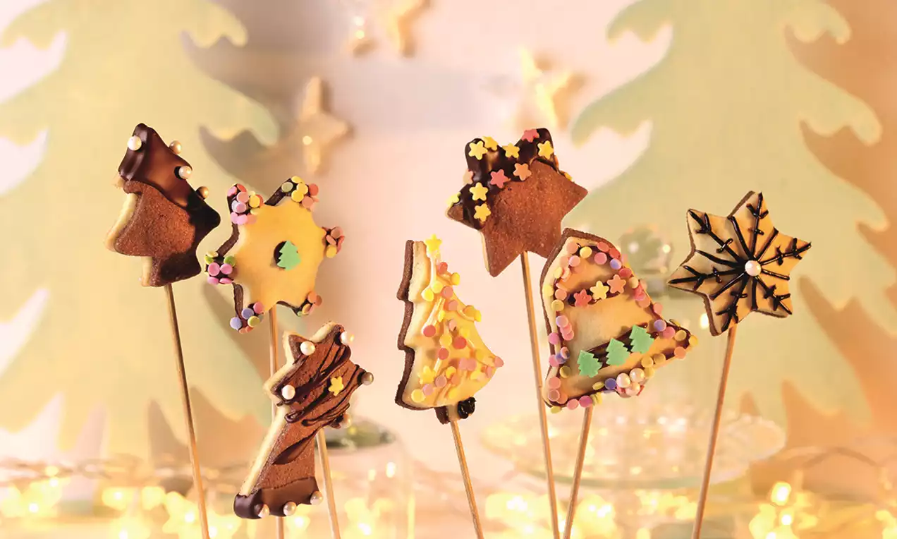 Božićni keksi na štapiću u raznim oblicima, ukrašeni šarenim čokoladnim preljevima, posipima i zvjezdicama na osvijetljenoj pozadini