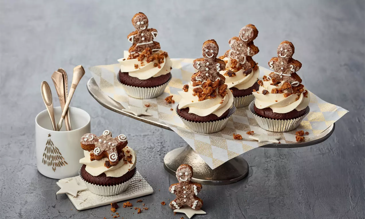 Čokoladni cupcakes s kremastim svijetlim šlagom na vrhu, ukrašeni smeđim medenjacima, mrvicama biskvita i posluženi na stalku s uzorkom.