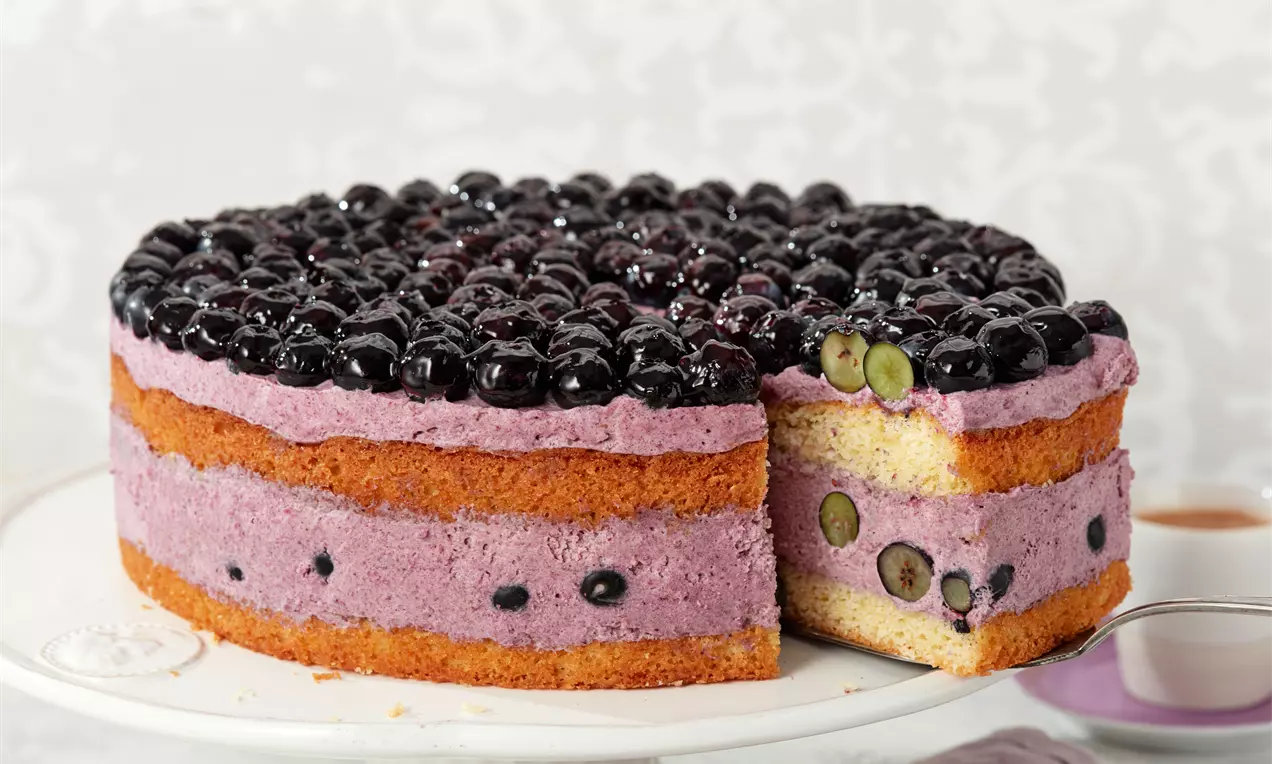 Torta okruglog oblika s dvije zlatnosmeđe biskvitne kore, slojevima ljubičaste kreme i glazurom od sjajnih cijelih borovnica